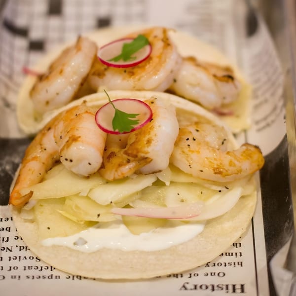 Two shrimp tacos with white sauce, shredded cabbage, and radish slices on newspaper-lined tray.