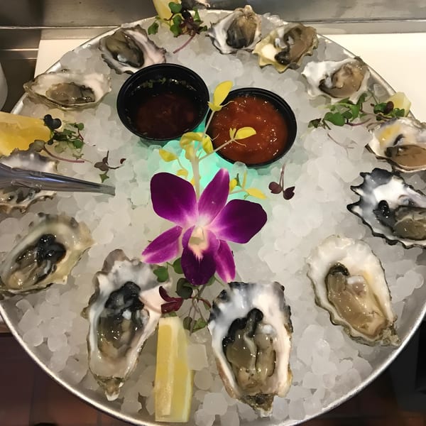 A dozen fresh oysters on ice with lemon, cocktail sauce, and a purple orchid.